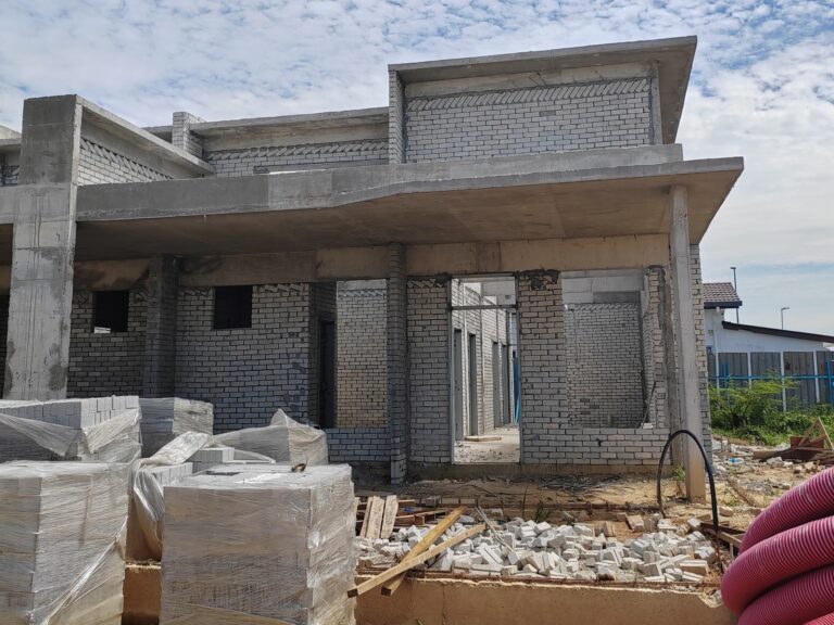 Foto sebenar tapak: fasad rumah setingkat dalam pembinaan dengan dinding bata kelabu, rasuk konkrit dan anjung kereta
