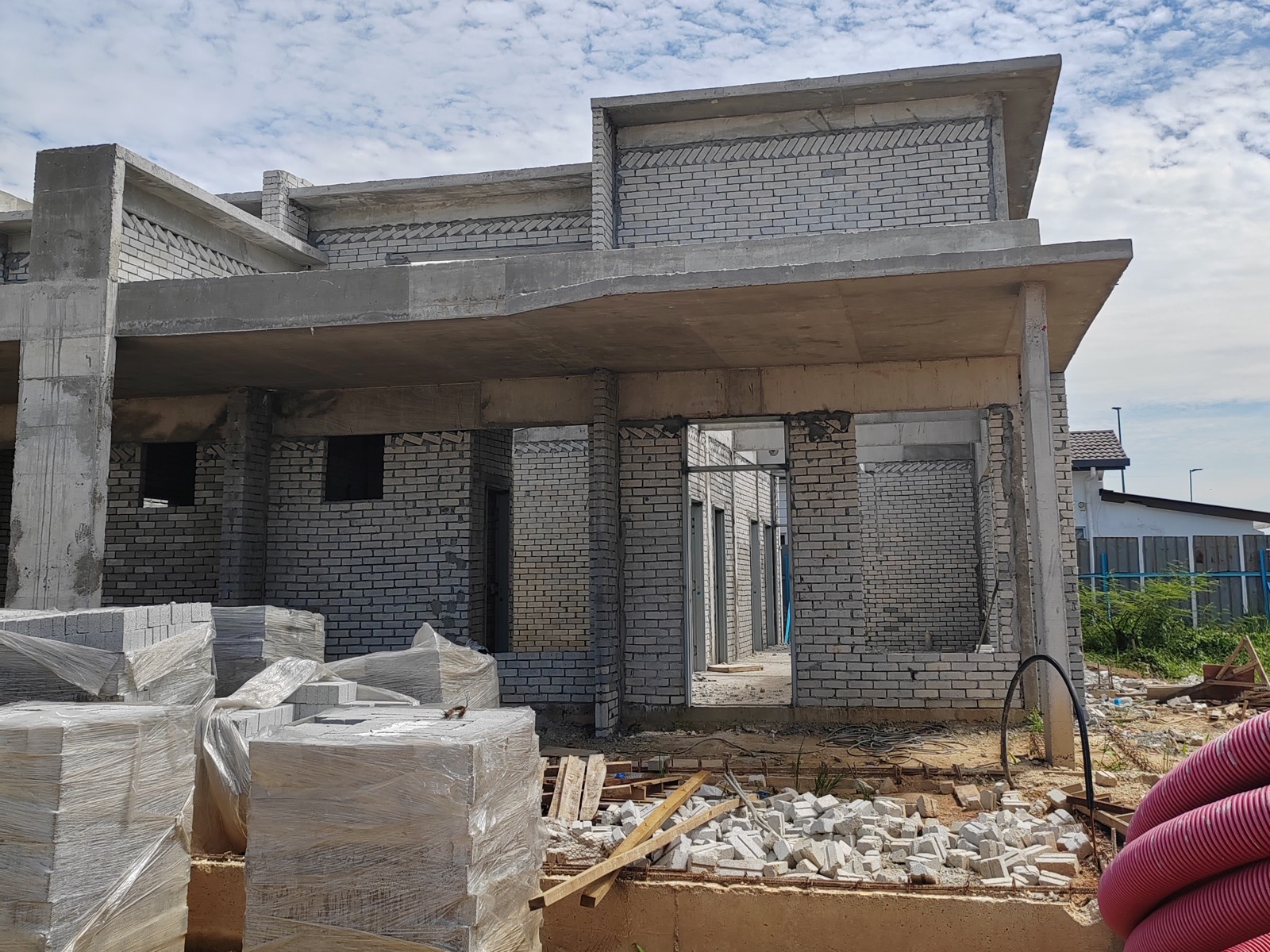 Foto sebenar tapak: fasad rumah setingkat dalam pembinaan dengan dinding bata kelabu, rasuk konkrit dan anjung kereta