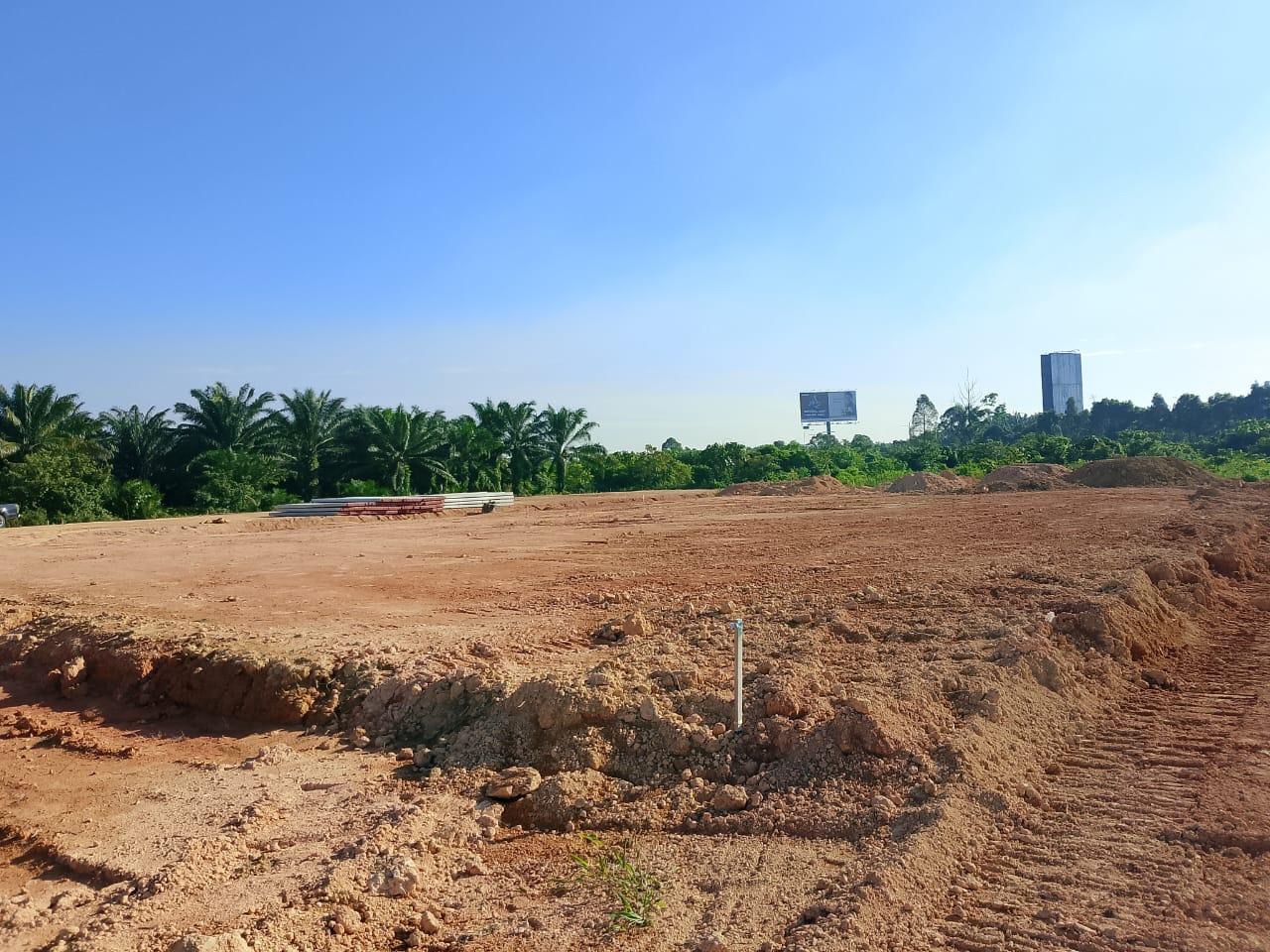 Foto tapak projek yang telah diratakan dengan tanah merah, pokok kelapa sawit di latar belakang