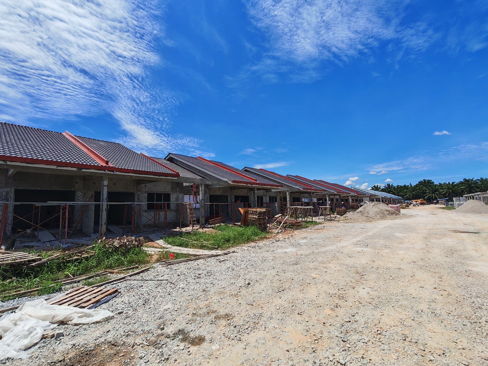 Foto sebenar jalan tanah di tapak projek dengan deretan rumah teres satu tingkat berbumbung merah di sebelah kiri
