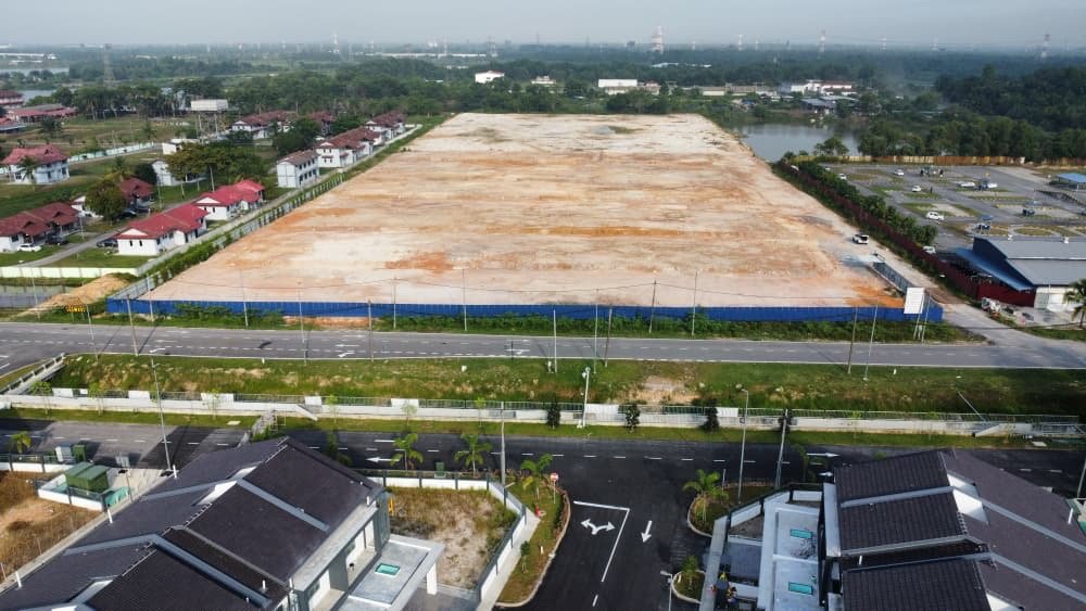 Foto dron sebenar tapak Taman Warisan Hijauan: tanah lapang yang telah dibersihkan dan berpagar biru di tepi jalan, dengan tasik dan rumah kediaman di sekeliling